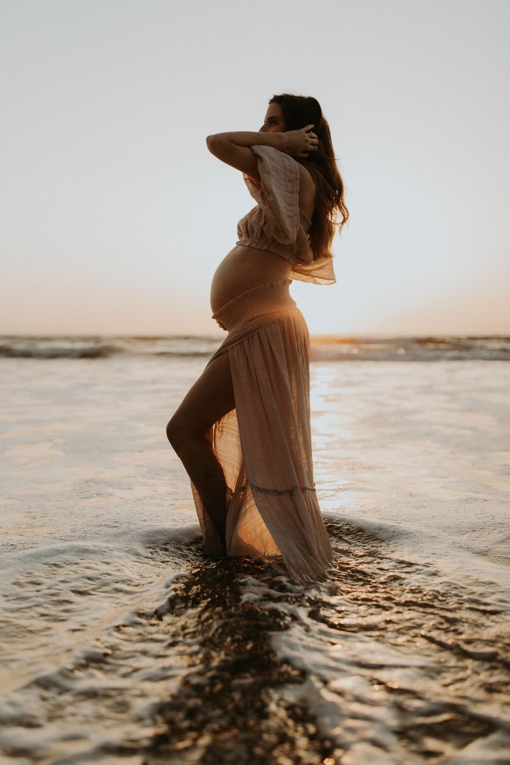 A person in a flowing dress stands in shallow ocean water at sunset creating a dreamy silhouette effect.
