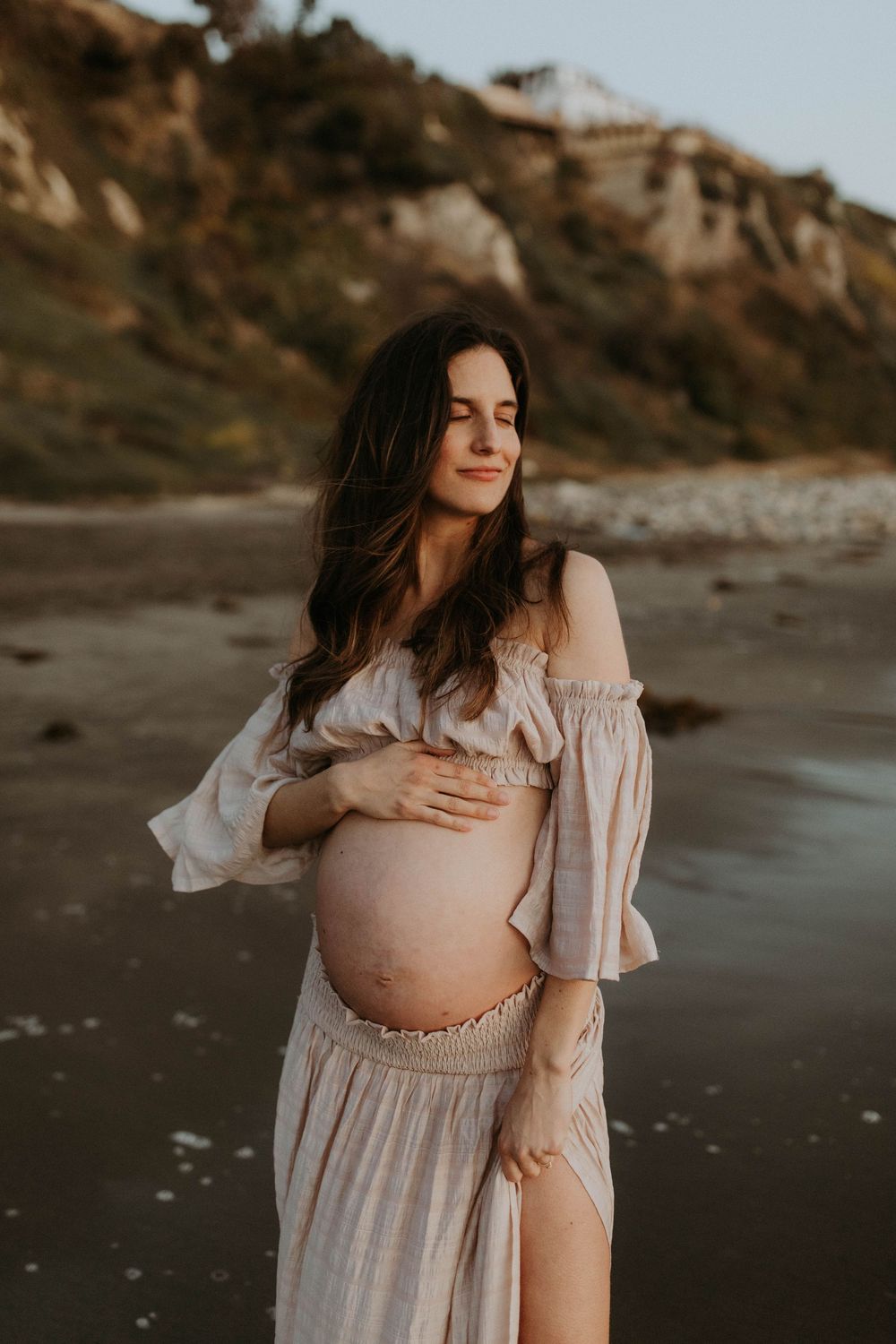 Pregnant woman in off-shoulder dress cradling baby bump on beach at golden hour.