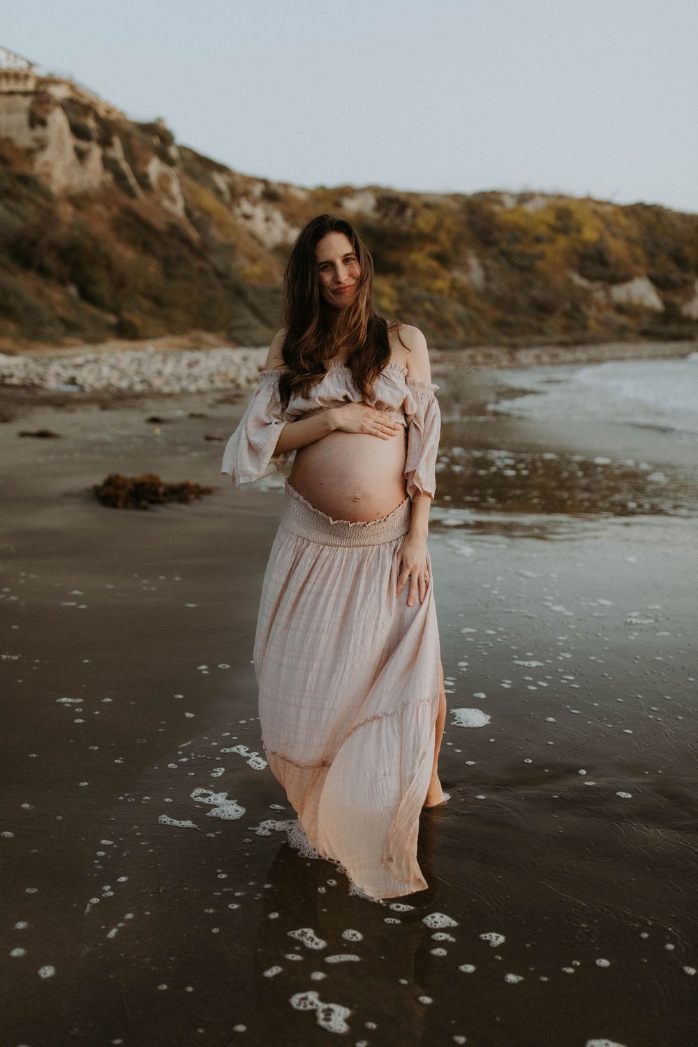 A pregnant woman in a flowing pink dress stands on a moody beach at sunset, cradling her baby bump.