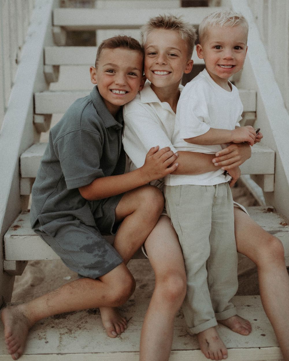 Children in gray shirts and beige overalls sit together on white steps sharing playful embraces and laughter.
