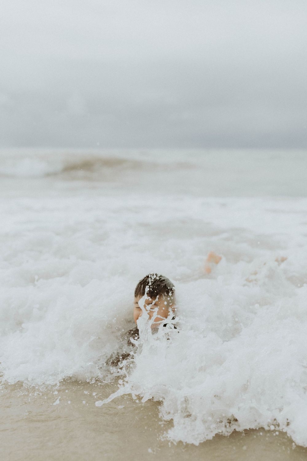 Child playing in ocean waves with white sea foam splashing around them on a cloudy beach day.