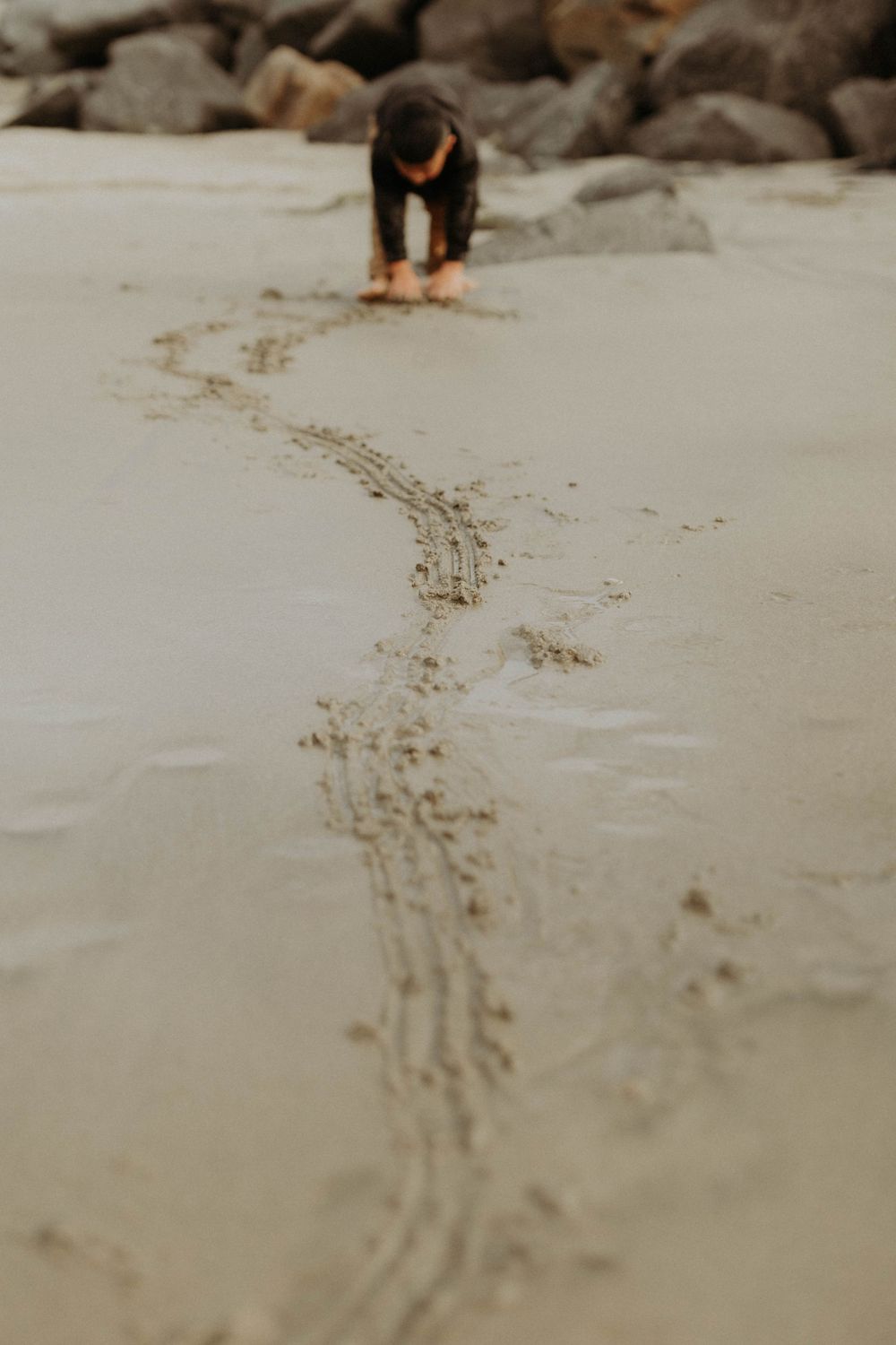 Child drawing a winding trail in wet beach sand near rocky shoreline.