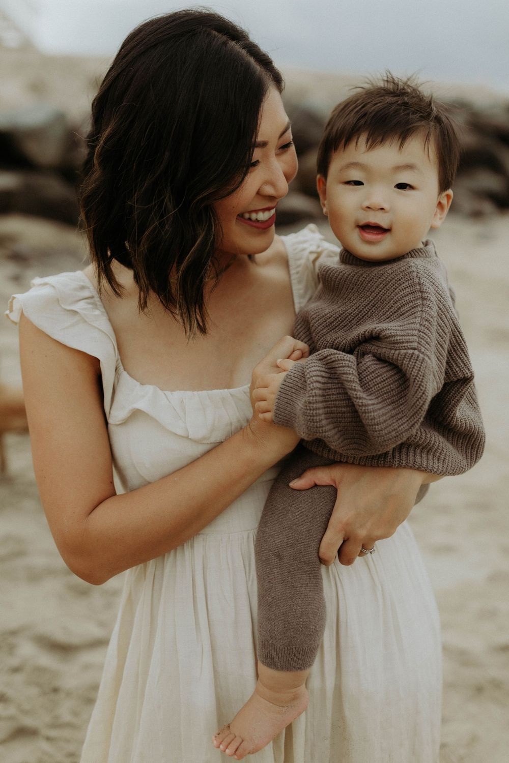 A parent and baby share a joyful moment together on the beach, wearing white dress and brown sweater.