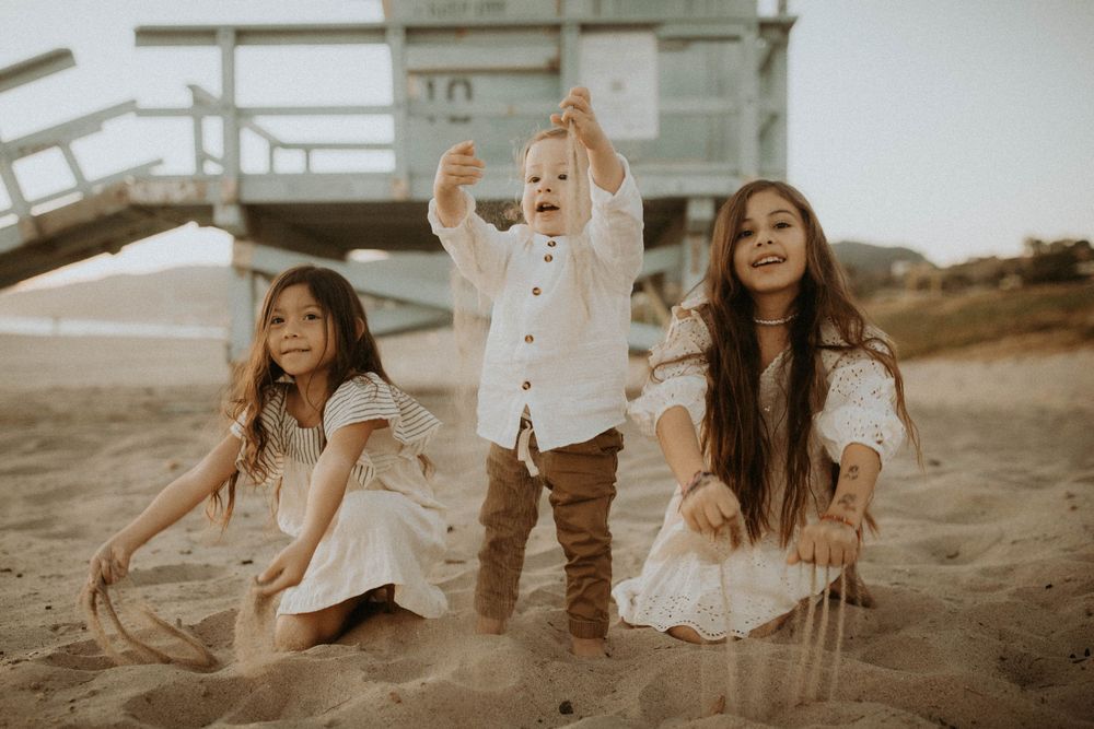 Three children play joyfully on a sandy beach near a lifeguard tower, two girls in white dresses flank a toddler boy.