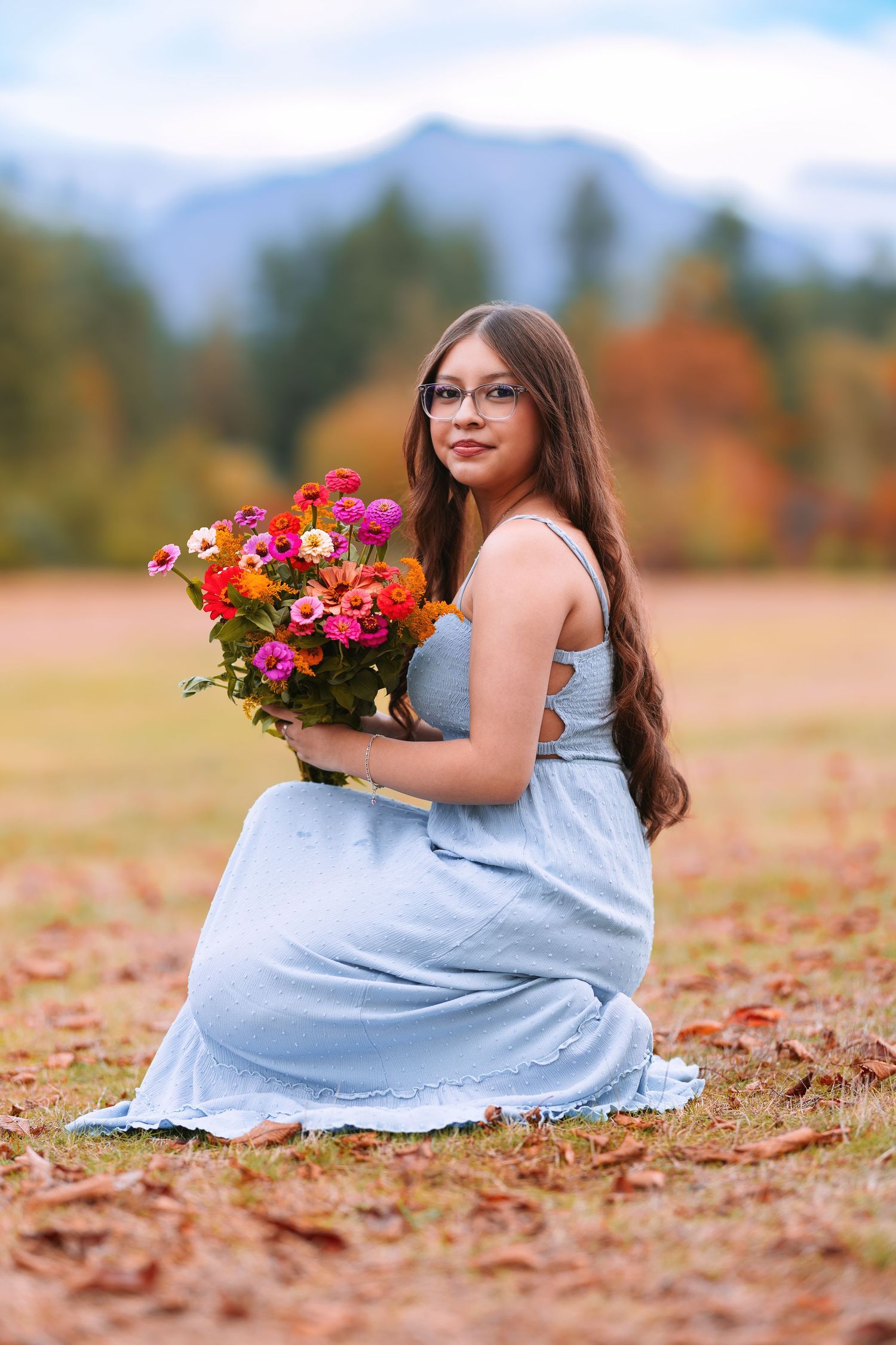 Young woman in blue dress holding colorful flower bouquet, sitting in autumn field with mountain backdrop.