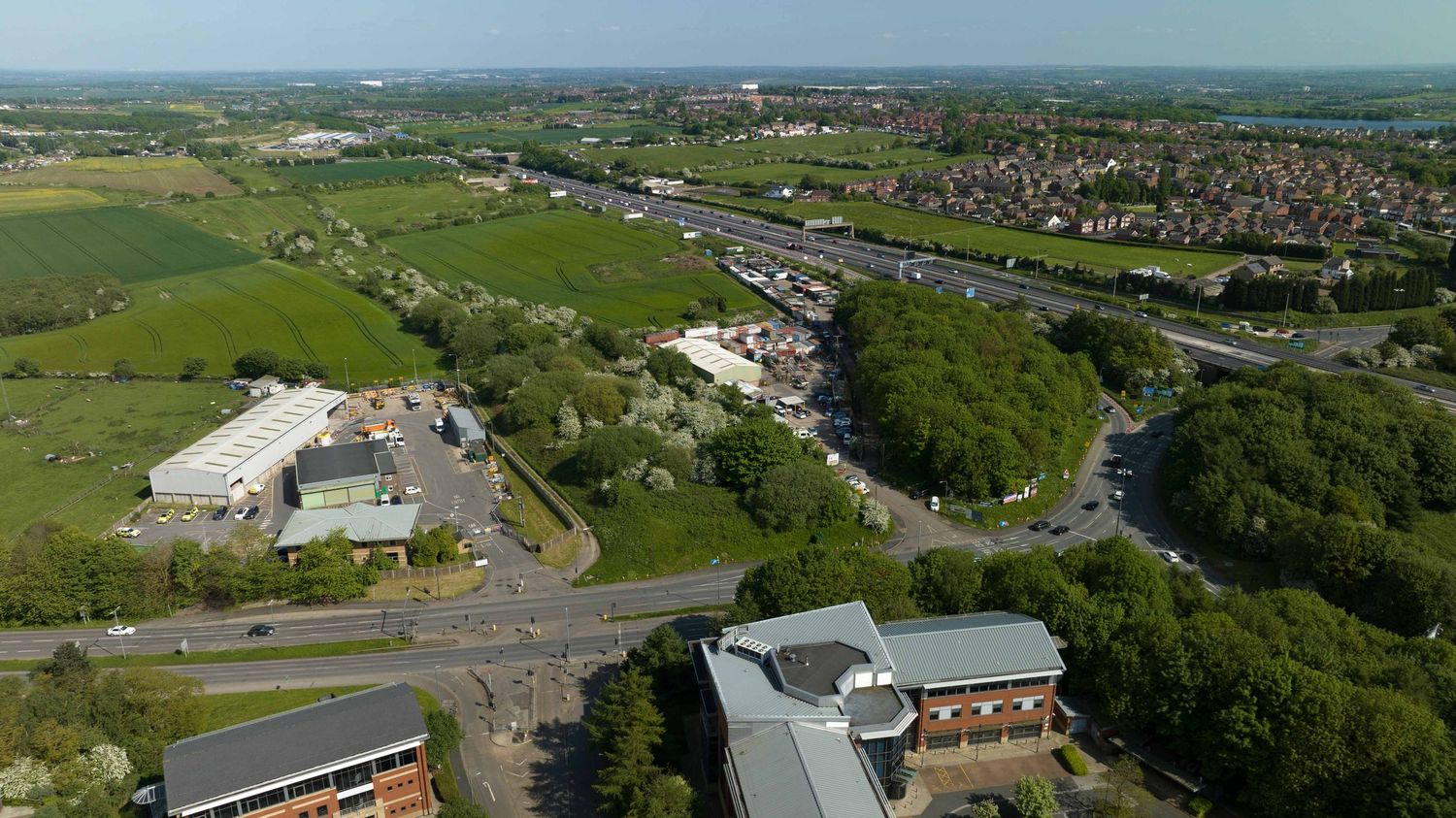 Aerial view of a UK town showing green fields, residential areas, a busy road, and commercial buildings on a clear day.