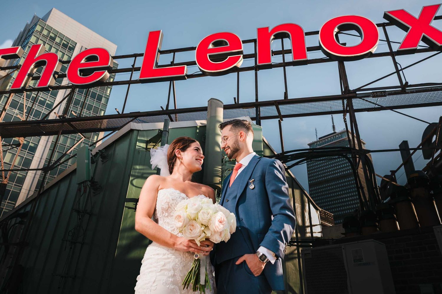 Bride and groom smiling at each other beneath The Lenox Hotel rooftop sign in Boston with city skyline views