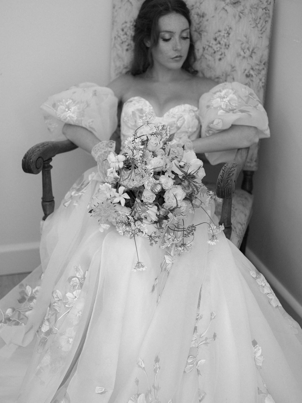 Bride in floral ball gown with puff sleeves holds cascading bouquet while seated in chair, black and white photo.