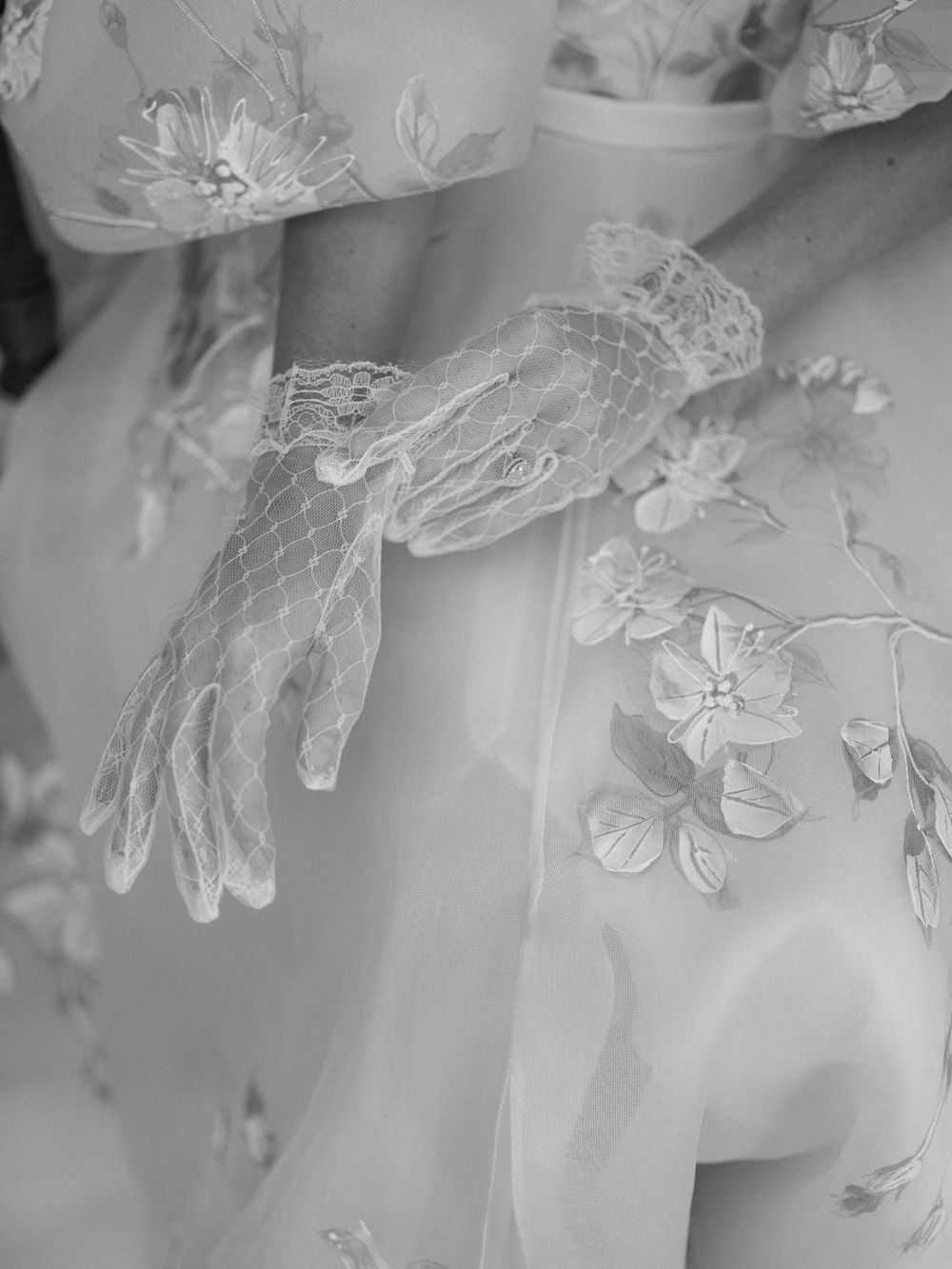 Close-up of a bride wearing delicate lace gloves and floral embroidered wedding dress in black and white.