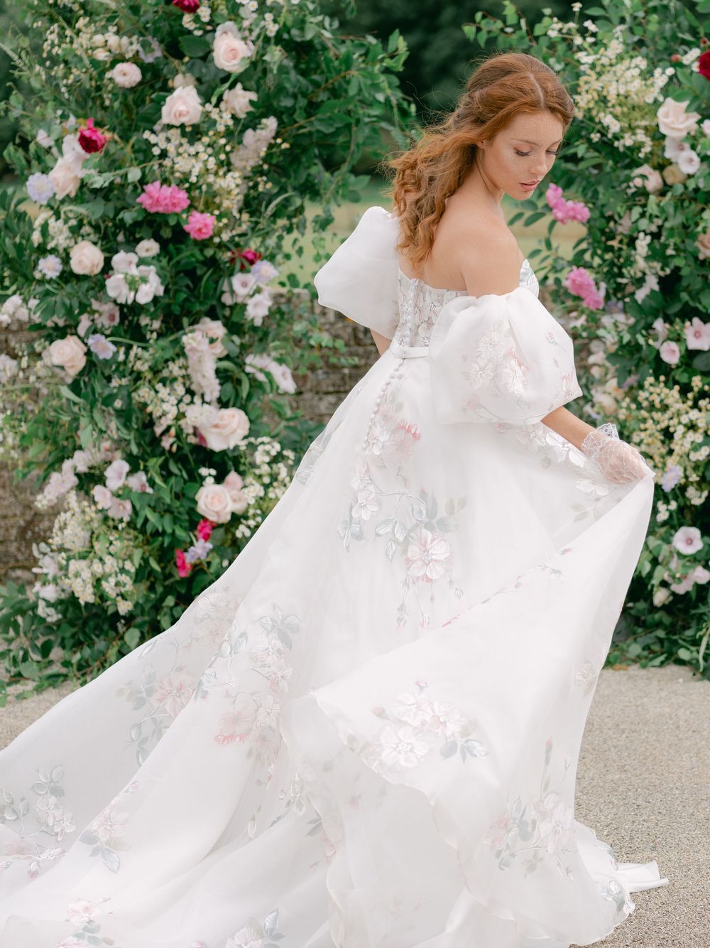 Bride in floral embroidered off-shoulder white gown poses before lush pink rose garden backdrop.