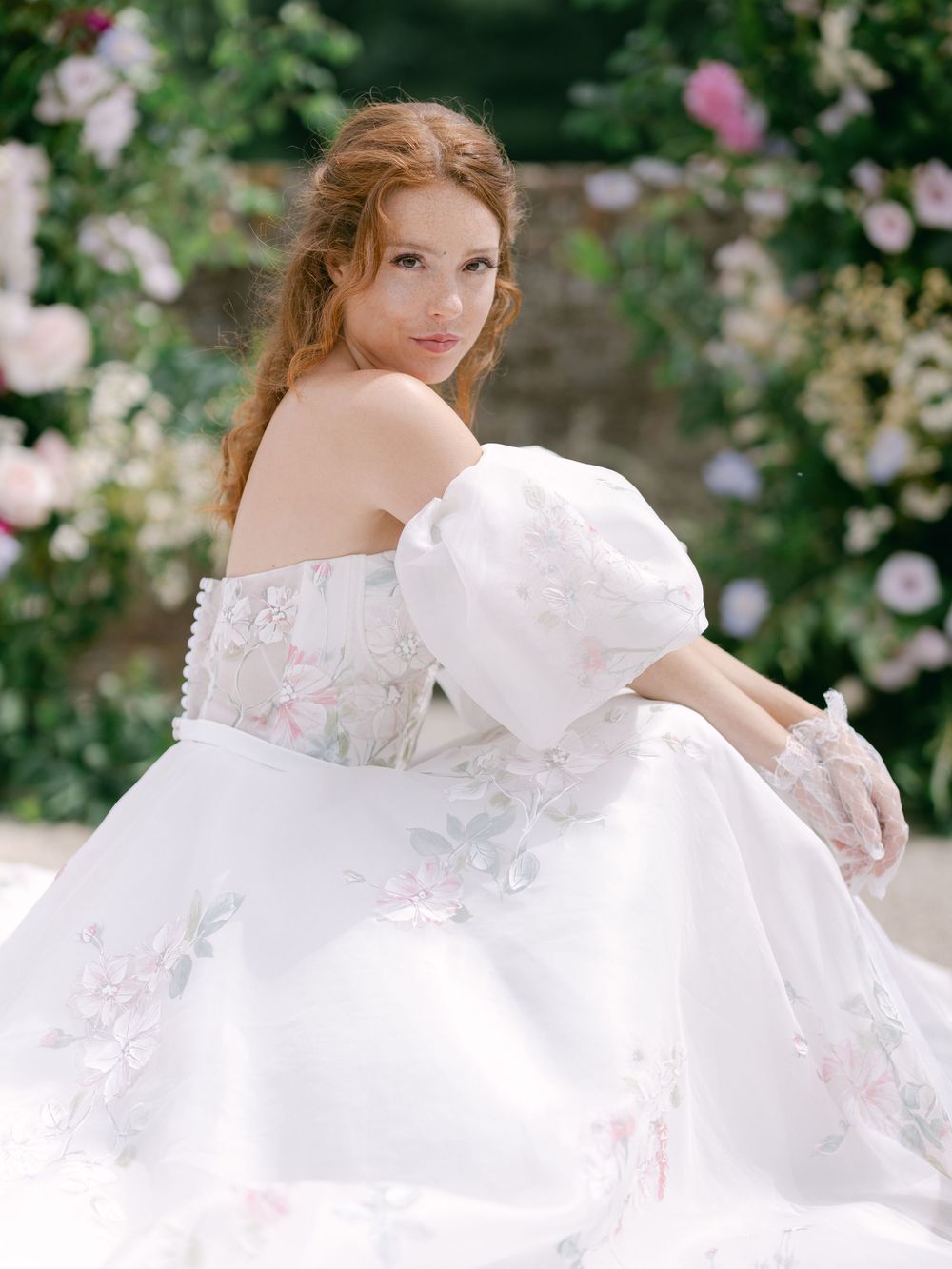Bride in floral off-shoulder ball gown with puff sleeves poses among pink roses in a lush garden setting.