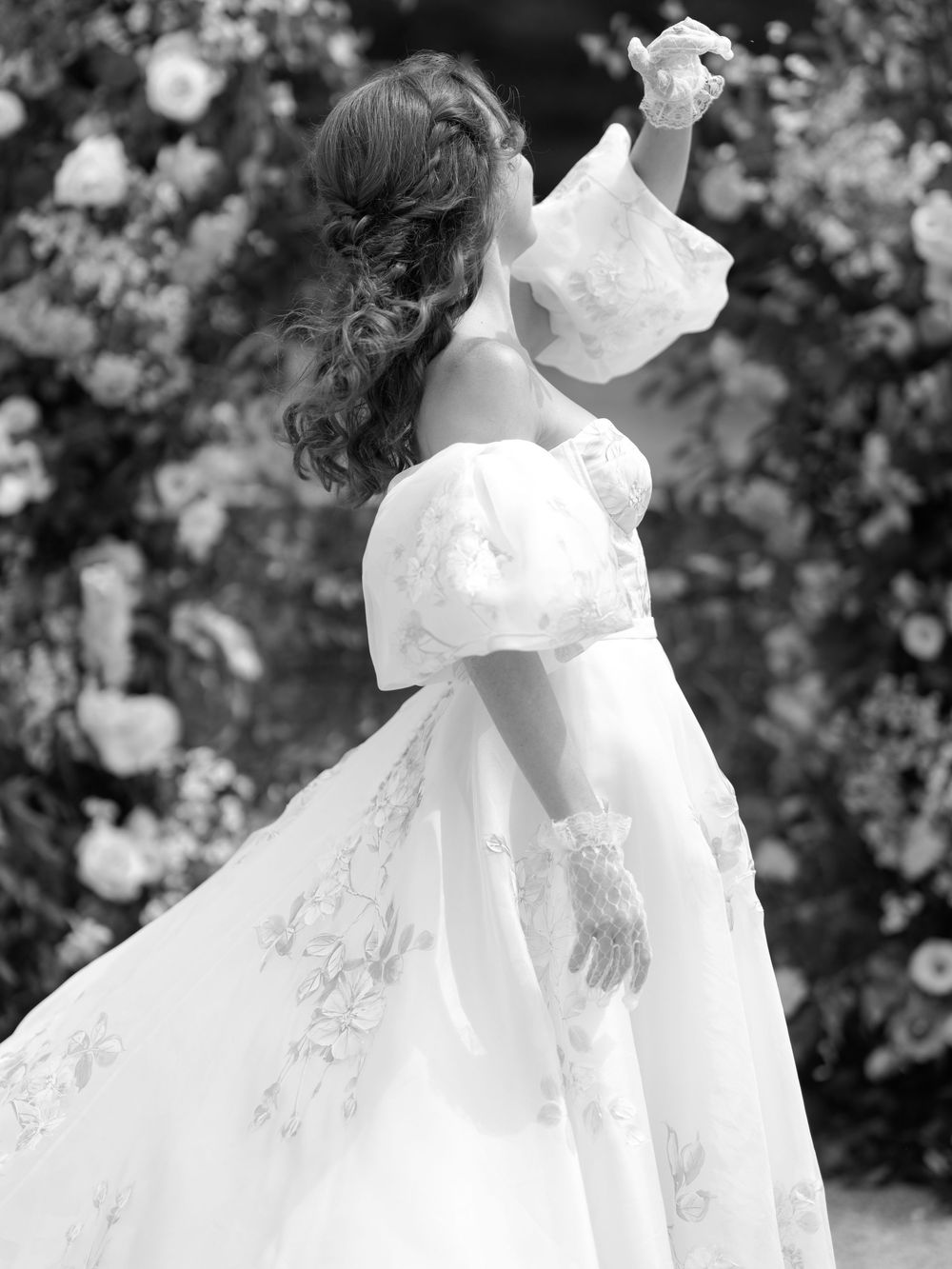 Black and white photo of a bride in an elegant off-shoulder gown with puff sleeves and floral details, posing by a floral backdrop.
