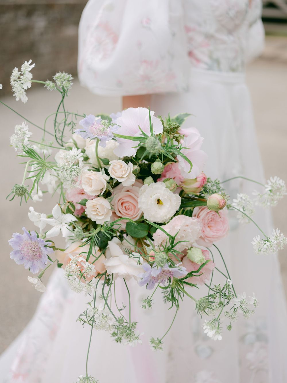Romantic bridal bouquet with blush pink roses, white ranunculus, lavender scabiosa, and wildflowers held by bride in floral gown.