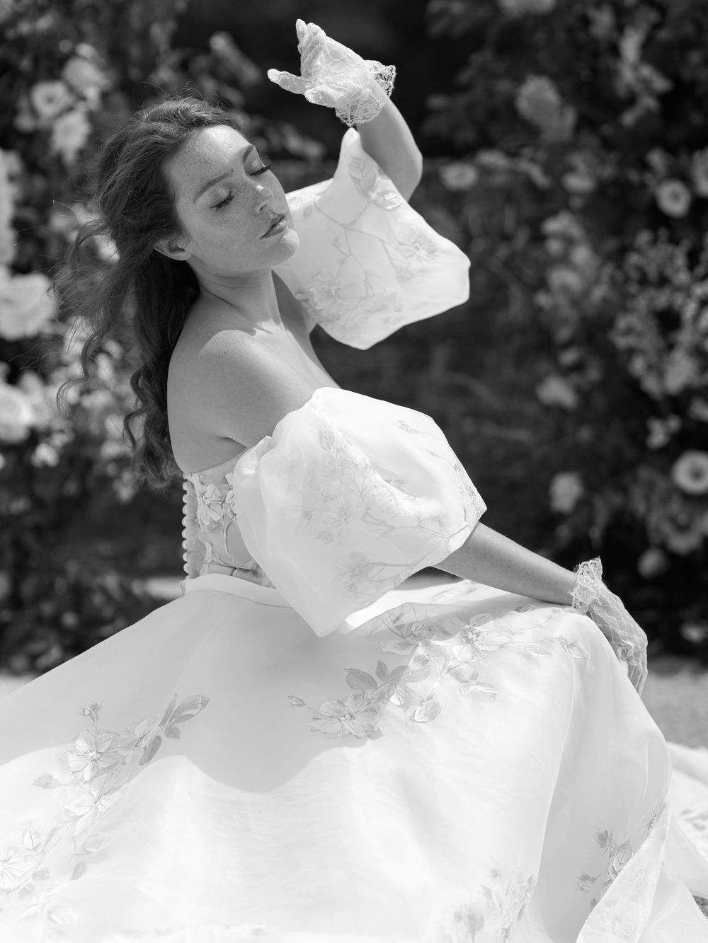 Black and white photo of bride in off-shoulder floral ballgown with puff sleeves and lace gloves in garden setting.