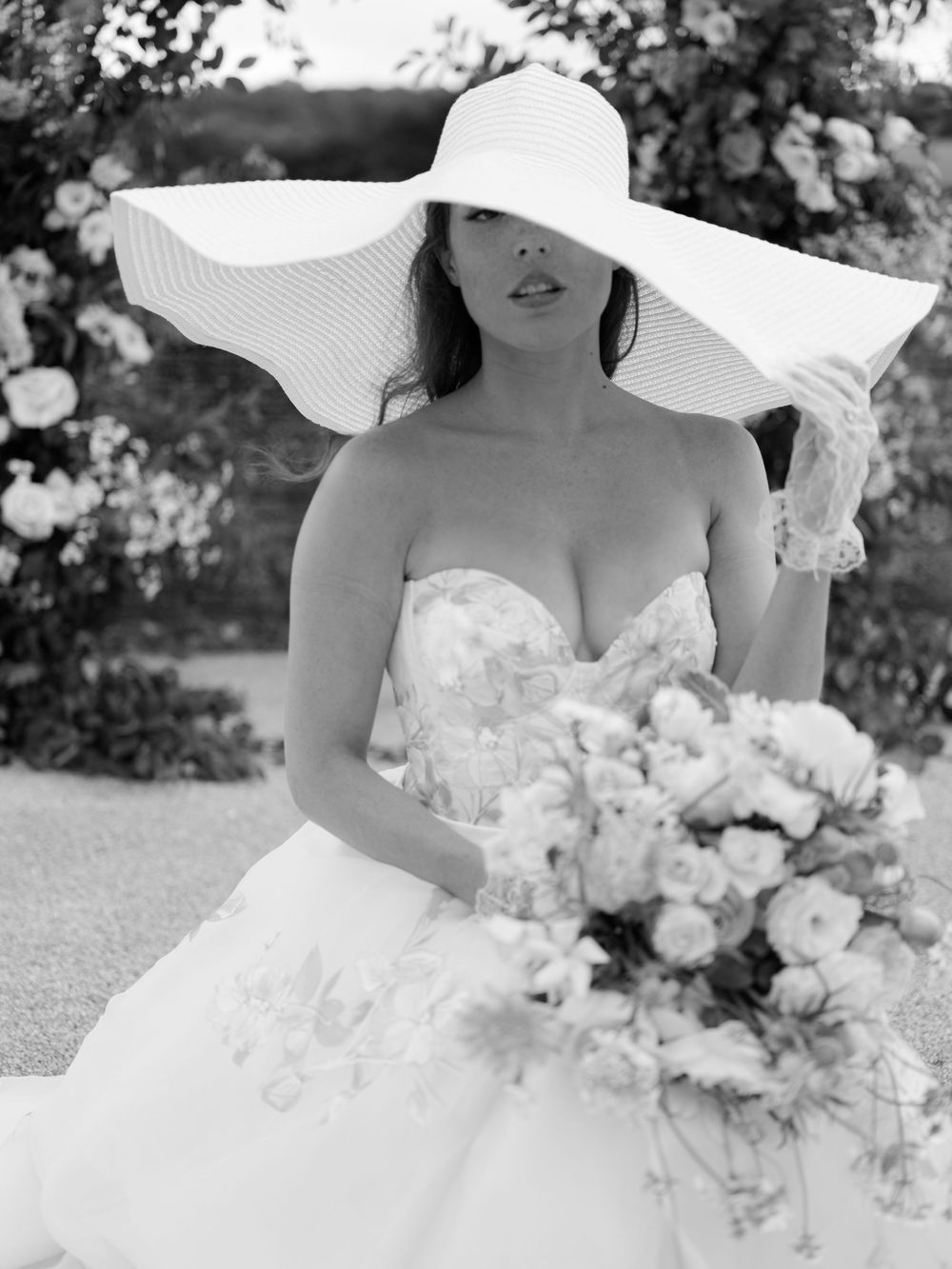 Bride in strapless floral gown and wide-brim white hat holding bouquet, black and white wedding photo.