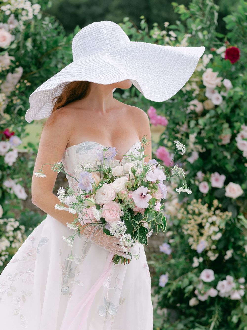 Elegant bride in strapless floral gown and wide-brim white hat holding pastel bouquet in lush garden.