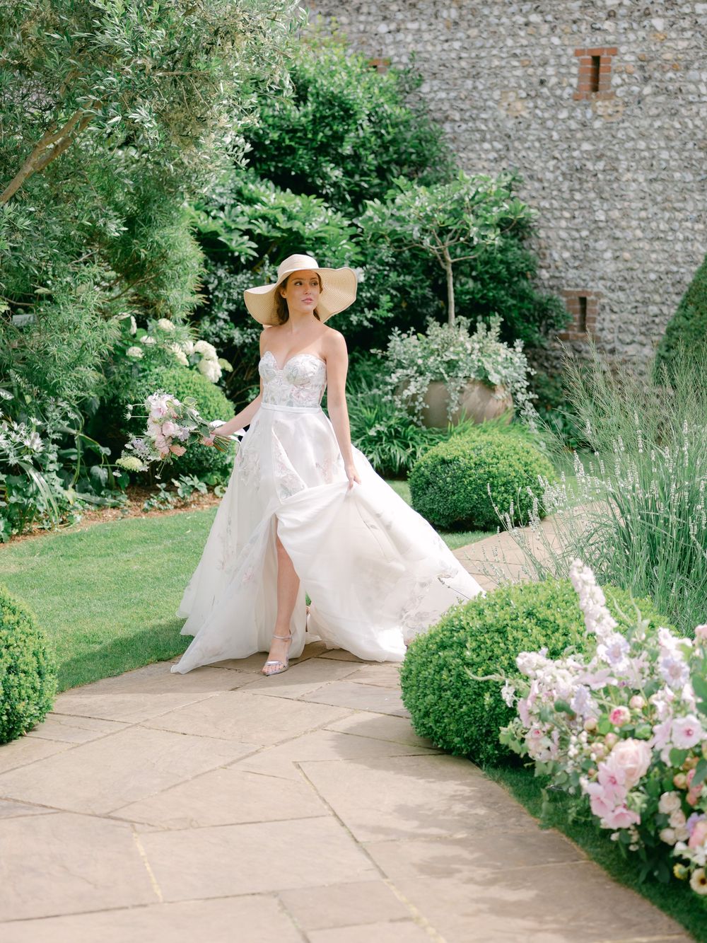 Bride in flowing lace wedding dress and sun hat walking through manicured English garden courtyard.