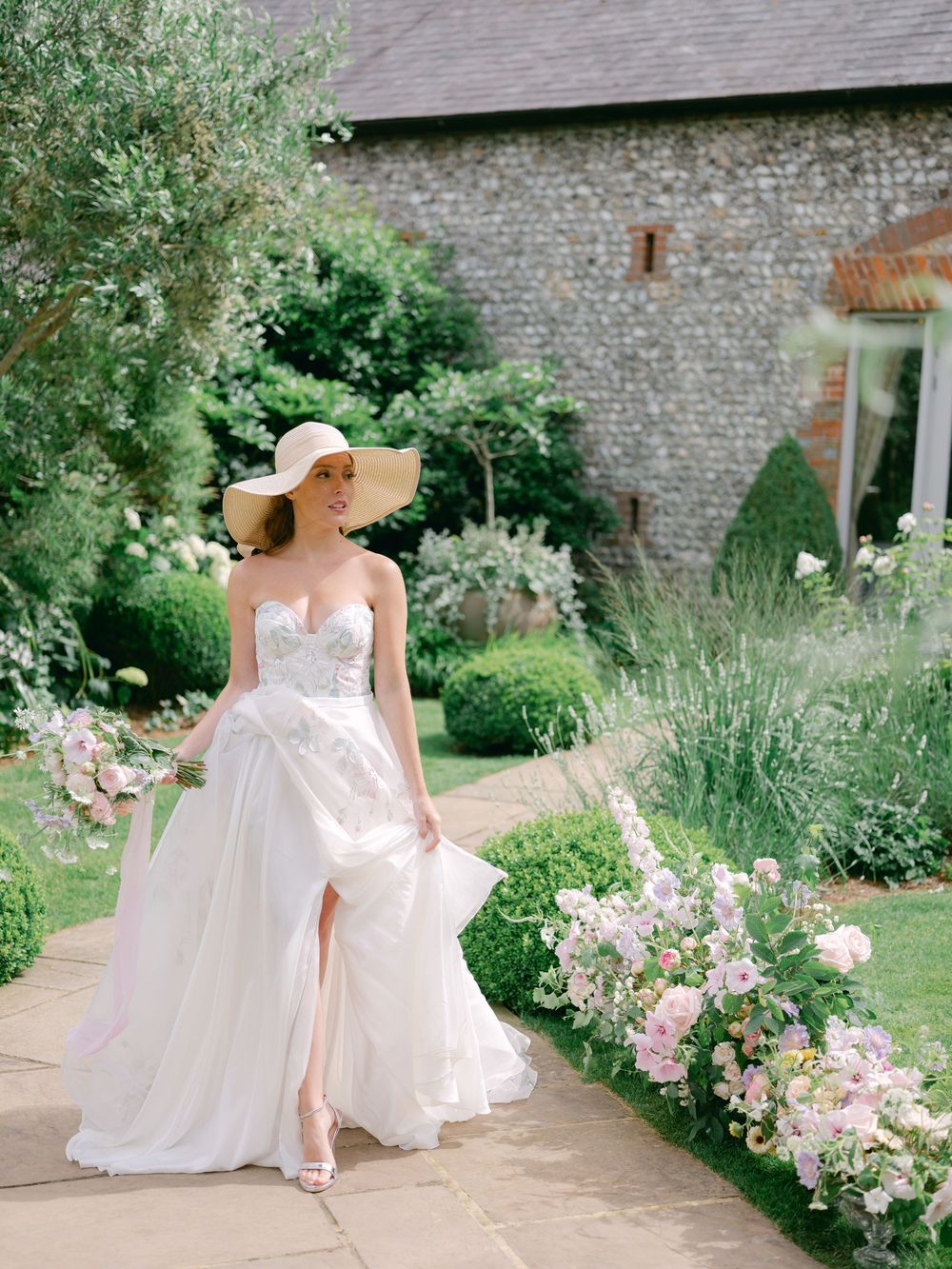 Bride in strapless lace wedding gown with thigh slit and wide-brim sun hat walks through an English garden.