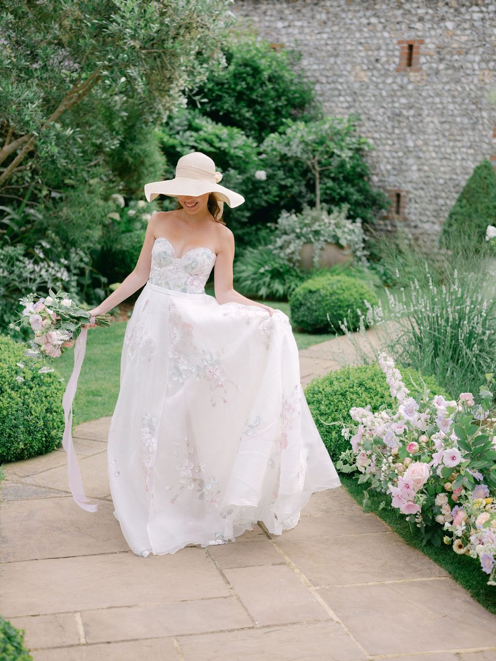 Bride in floral gown and wide-brim hat walks through lush garden path surrounded by pink blooms.