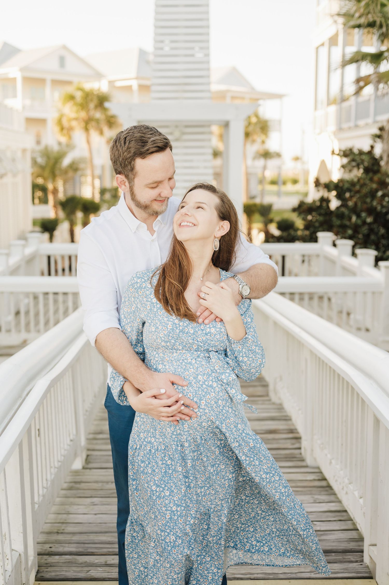 A romantic couple embraces on a white beach boardwalk while wearing coordinated blue outfits at sunset.