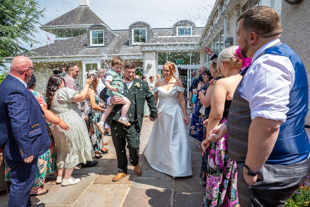 Newlyweds exit venue as guests shower them with confetti on a sunny wedding day.