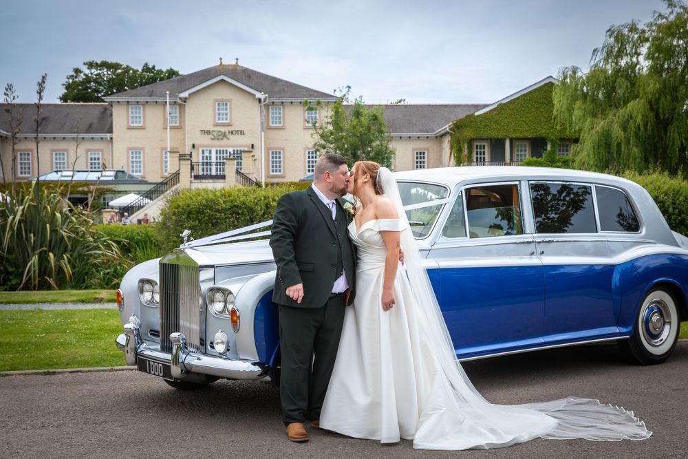 Bride and groom share a romantic kiss beside a classic blue and white Rolls-Royce wedding car.