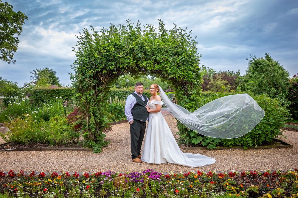 Bride's veil flows dramatically in the wind as couple poses under a garden arch.