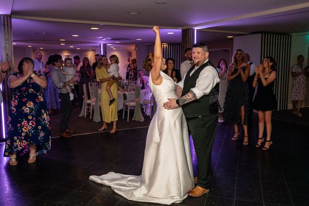Bride raises her arm joyfully during first dance surrounded by celebrating wedding guests.
