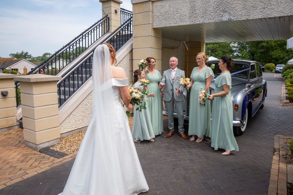 Bride in white gown approaches bridesmaids in sage green dresses beside a vintage blue car.