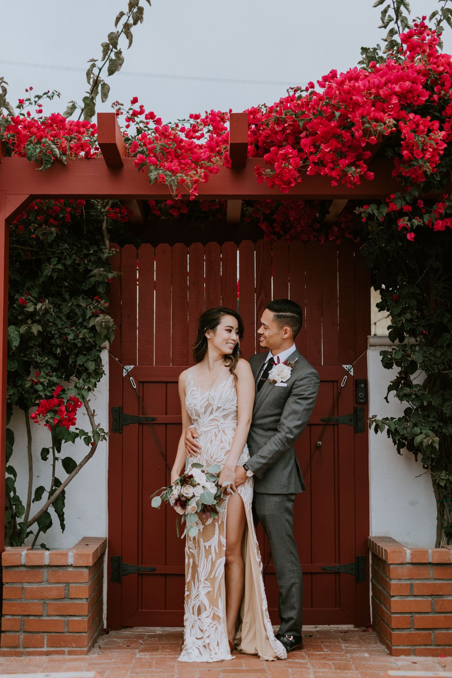 Elegant couple stands beneath cascading red bougainvillea at a charming Spanish-style doorway.