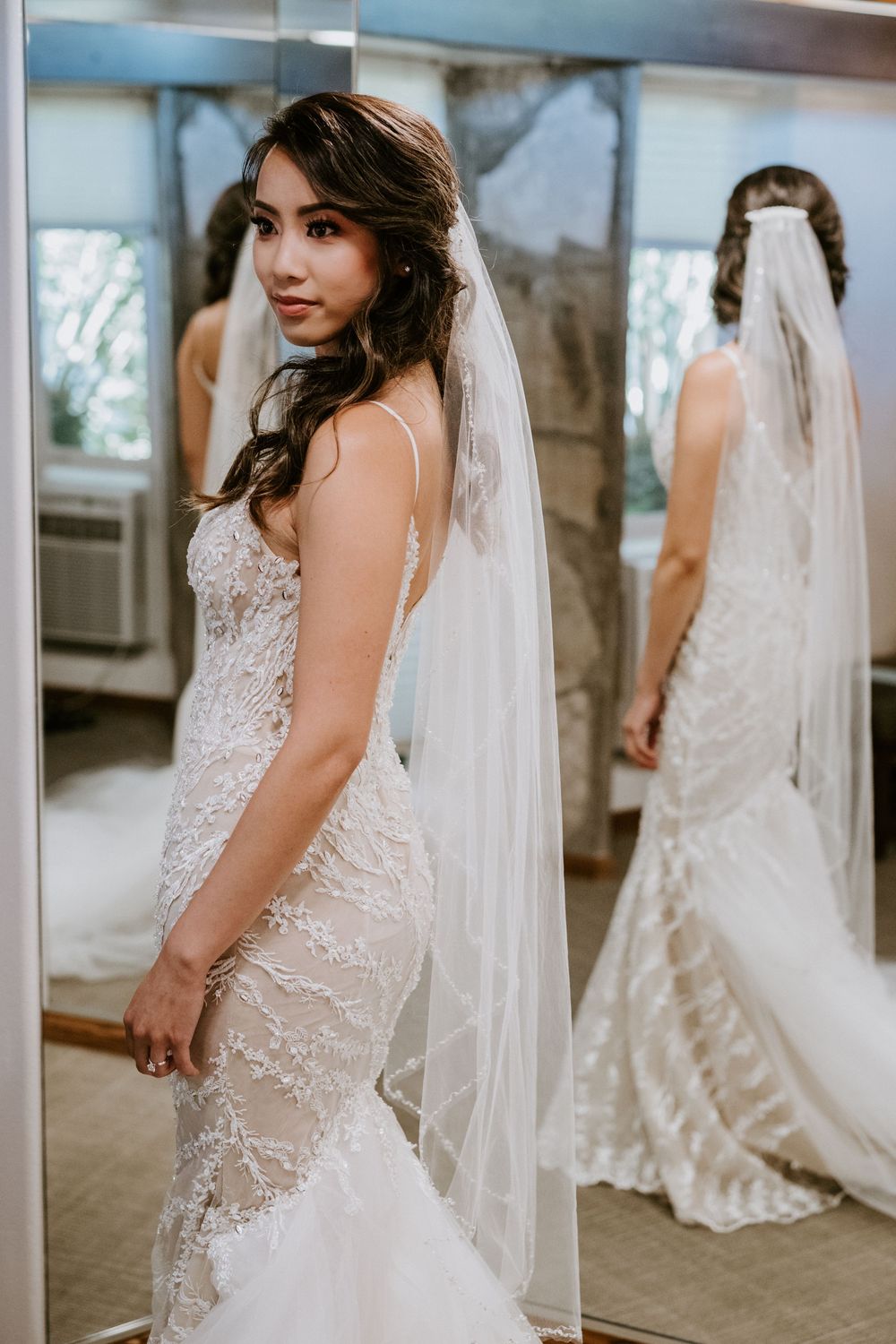 Bride poses in fitted lace mermaid wedding gown with veil in mirrored dressing room.