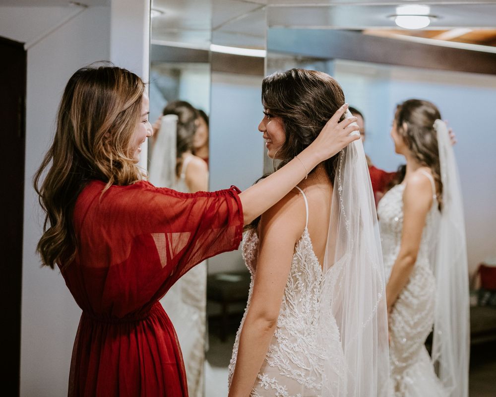 Bridesmaid in red dress adjusts brides veil in mirrored bridal suite before wedding.