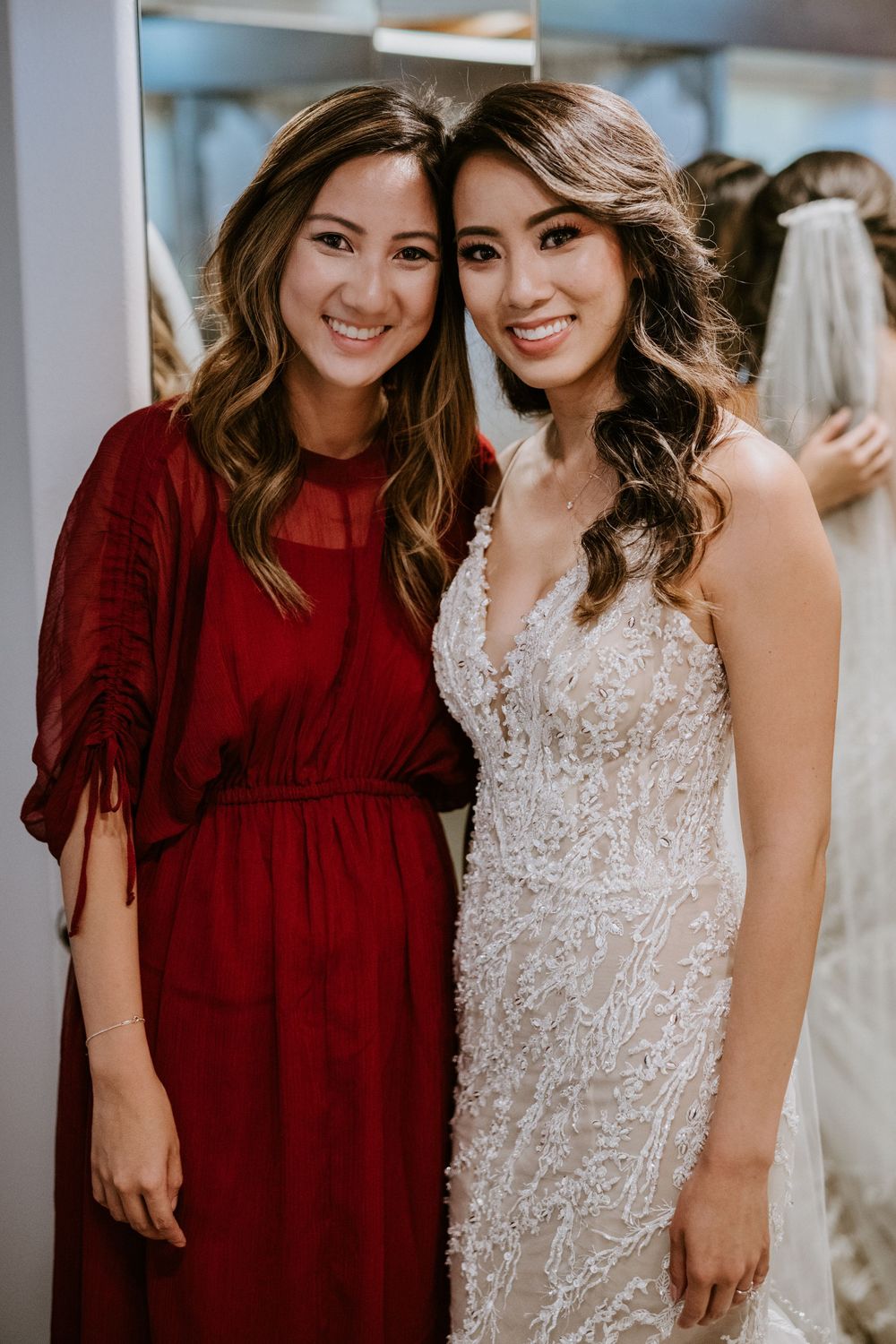 Bridesmaid in red dress poses with smiling bride in lace wedding gown before ceremony.