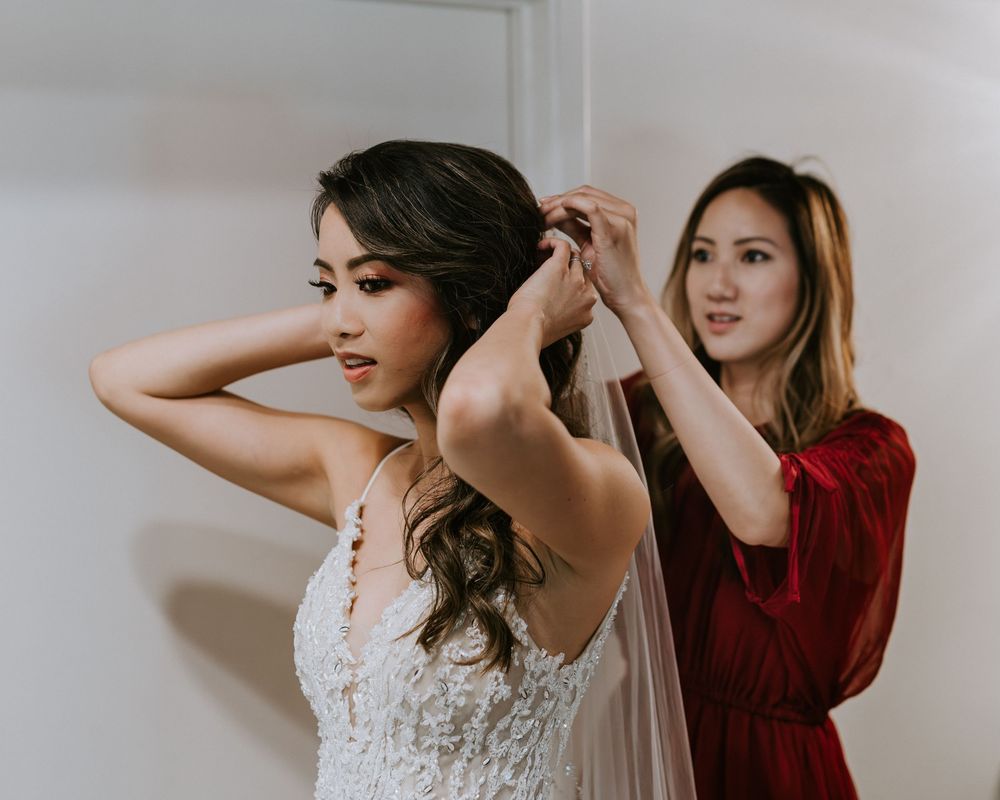 Bridesmaid in red dress helps bride with veil during wedding preparations.