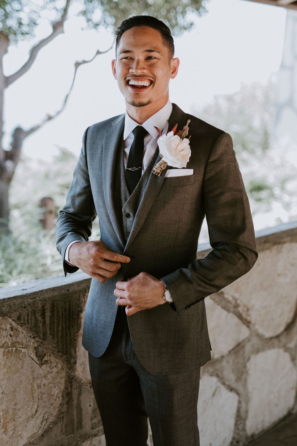 Dapper groom in charcoal three-piece suit with floral boutonniere smiles on wedding day.