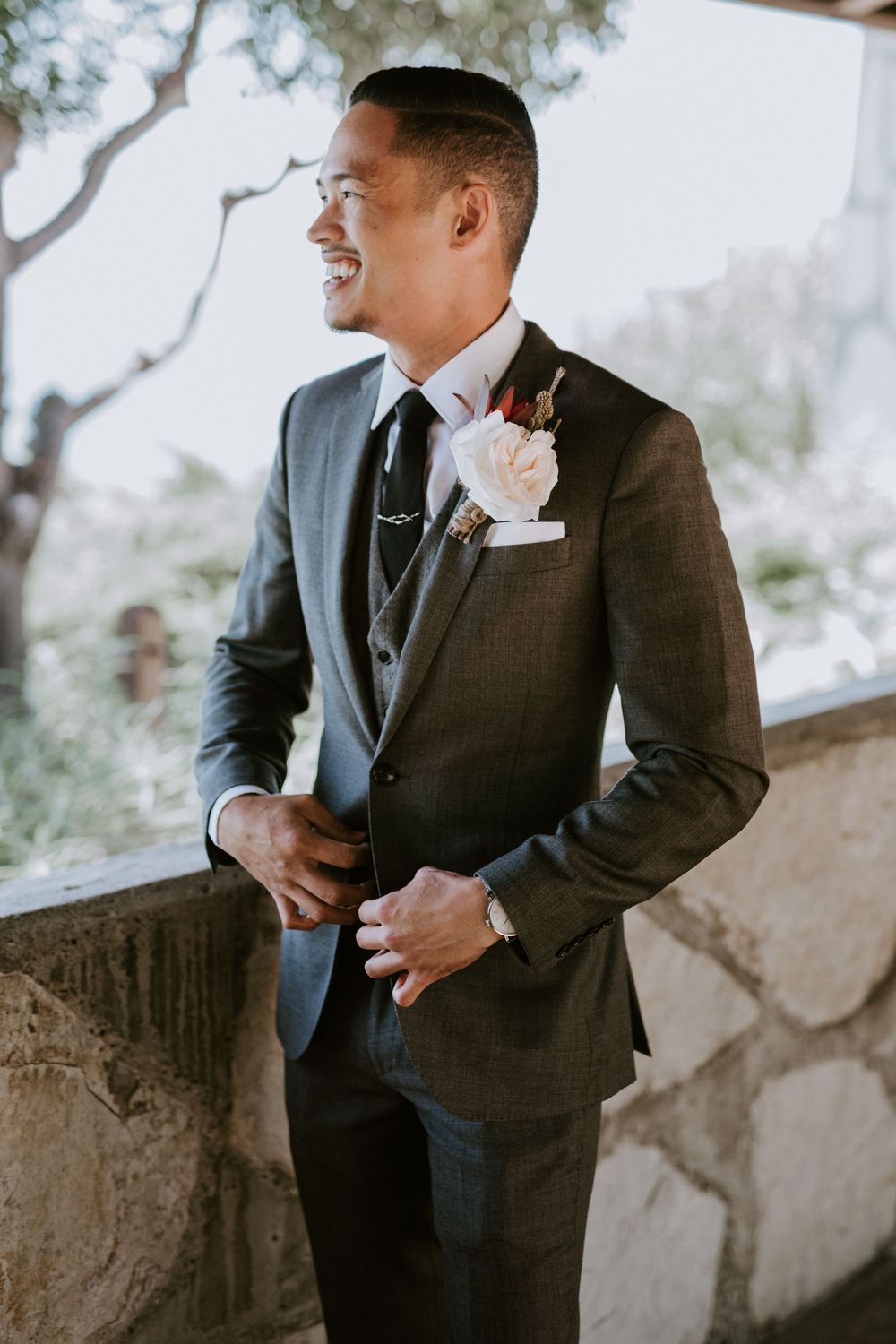Smiling groom in gray three-piece suit with boutonniere gazes outdoors on wedding day.