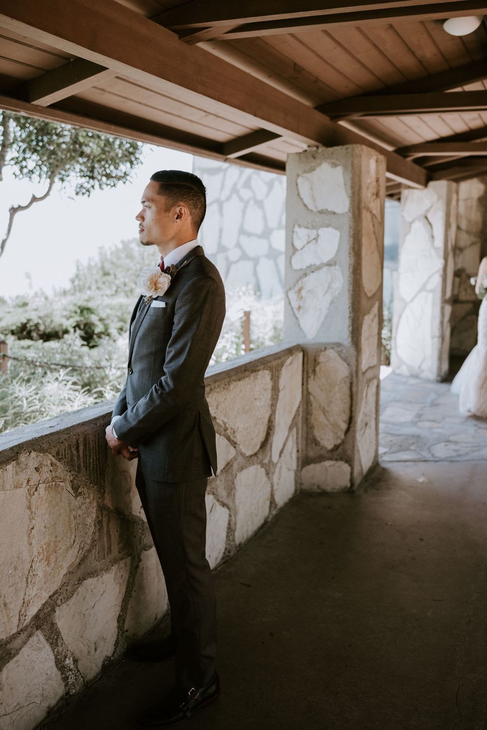 Groom in charcoal suit stands pensively on stone venue terrace before wedding ceremony.