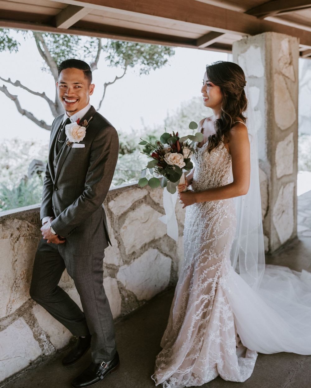 Smiling groom in gray suit stands beside bride in lace wedding gown holding bouquet at stone venue.