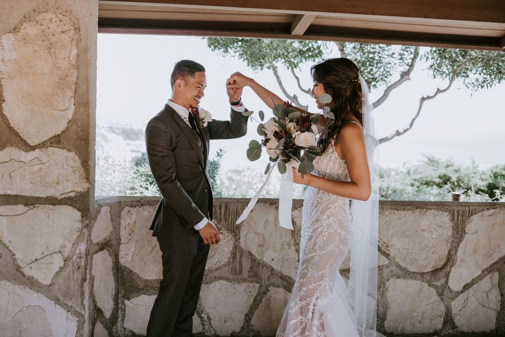 Groom and bride share a joyful first look moment, holding hands with floral bouquet at stone venue.