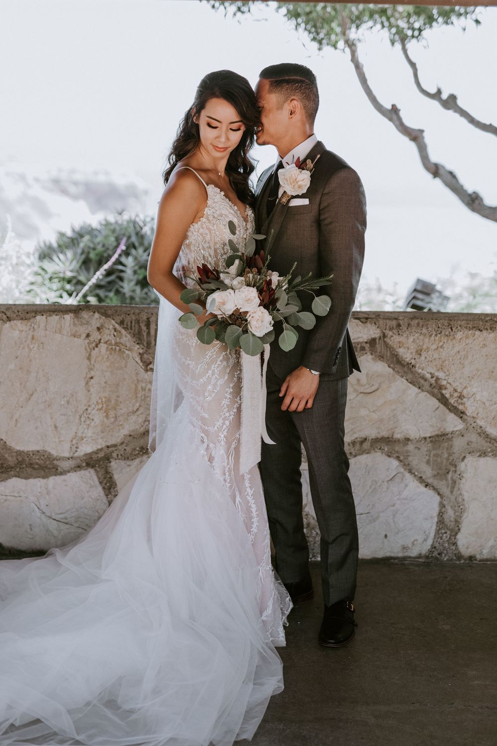 Groom tenderly kisses bride's temple as she holds lush bouquet, both in elegant wedding attire outdoors.