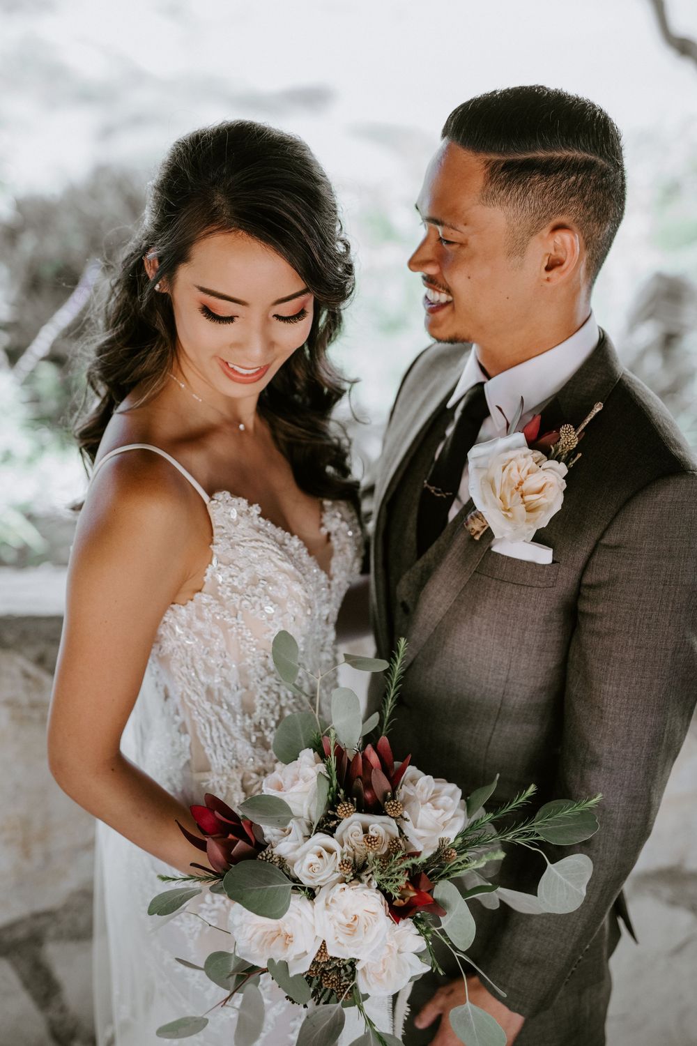 Bride and groom smile together, she holds white and red bouquet in lace gown, he wears gray suit.