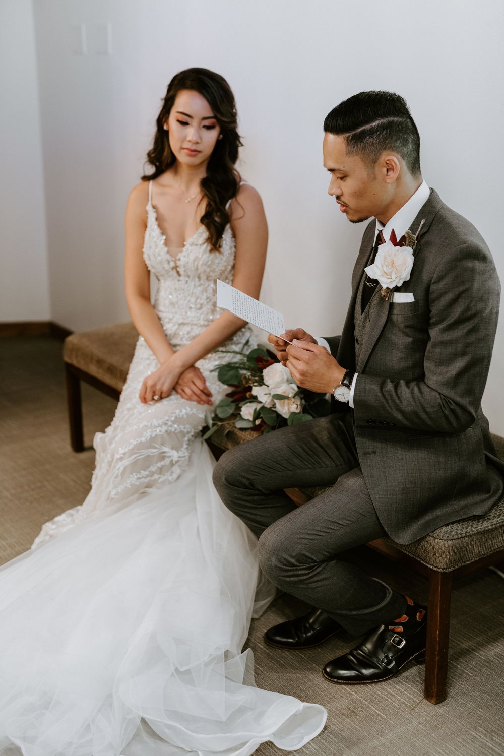 Bride and groom sit together reading vows, bride in lace gown holding bouquet in softly lit room.