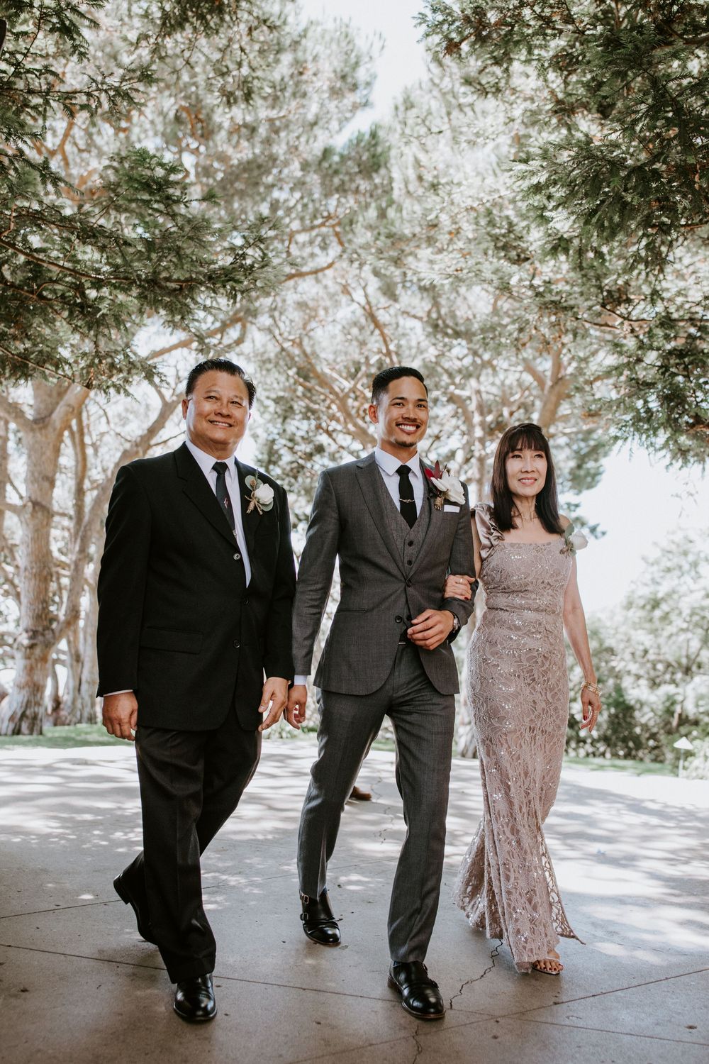 Smiling groom walks with parents outdoors under tall trees, all dressed in formal wedding attire.