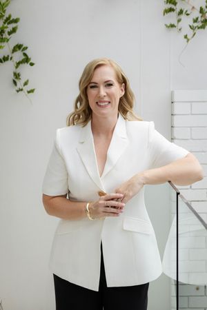 Business professional in white blazer posing confidently against white brick wall with glass elements.