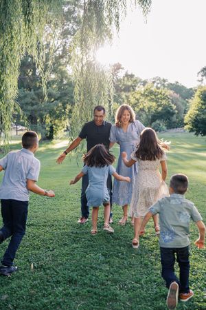 Family members run and play together on a grassy hill under willow trees in a summer park setting.