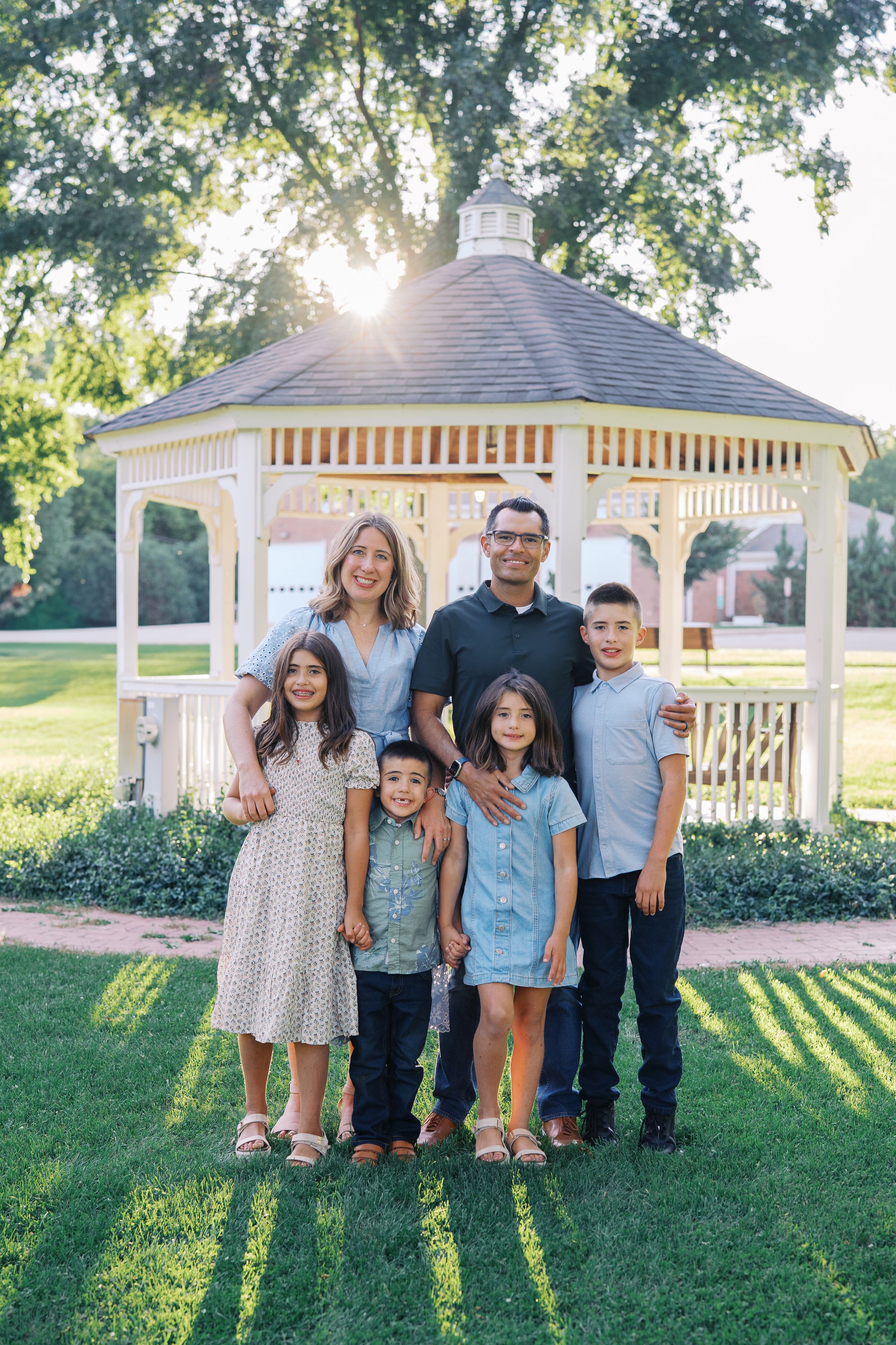 A family poses together near a white gazebo in a park setting during golden hour.