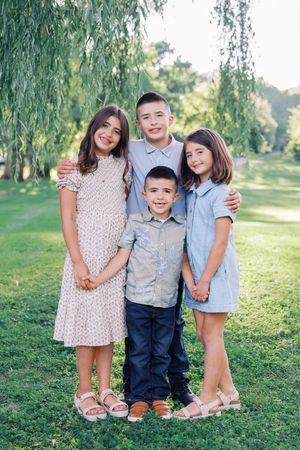 Three siblings stand together on grass under a weeping willow tree in coordinated blue and pink outfits.