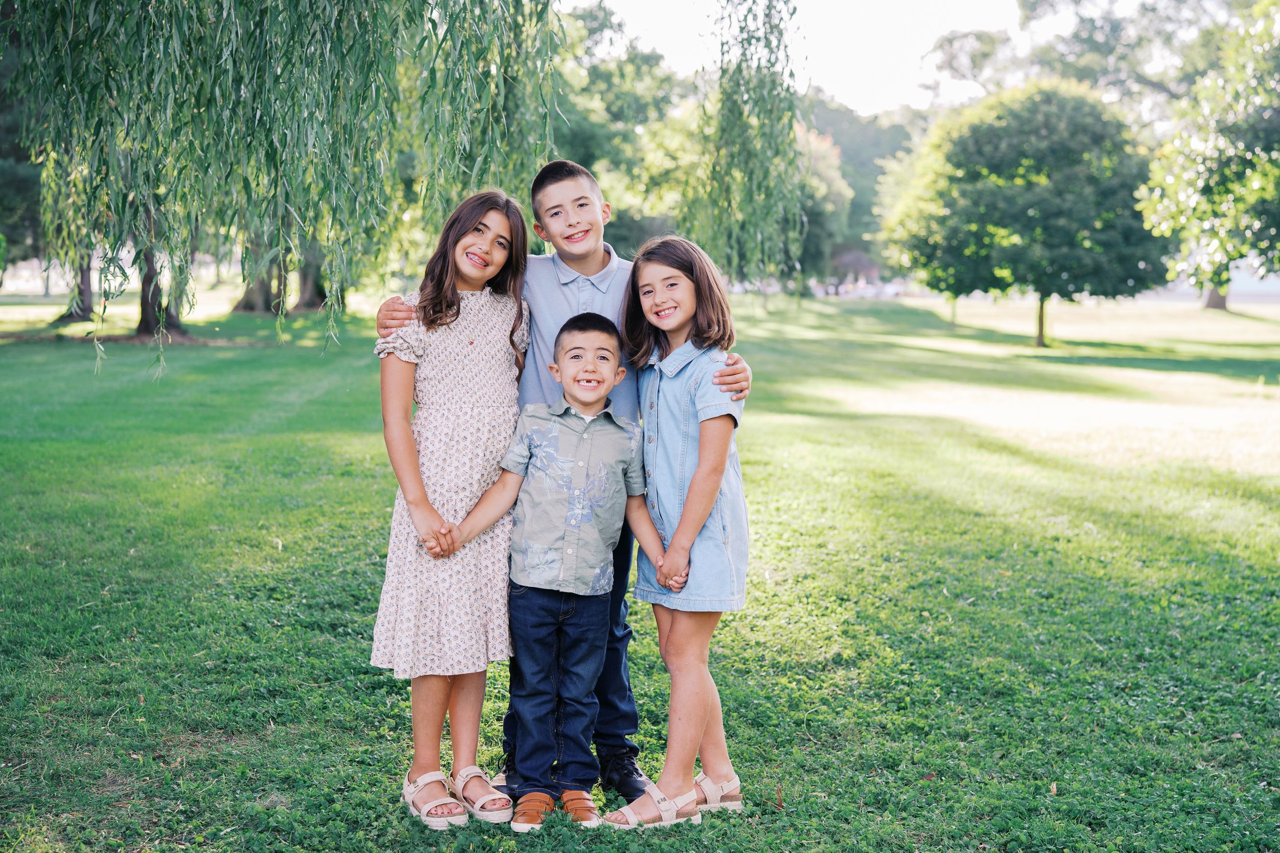 Three children pose playfully under drooping willow branches on a sunny day in a park.