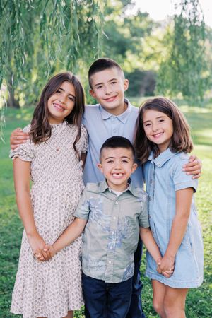 A casual outdoor family portrait series with children and adults posing together under green trees in natural lighting.