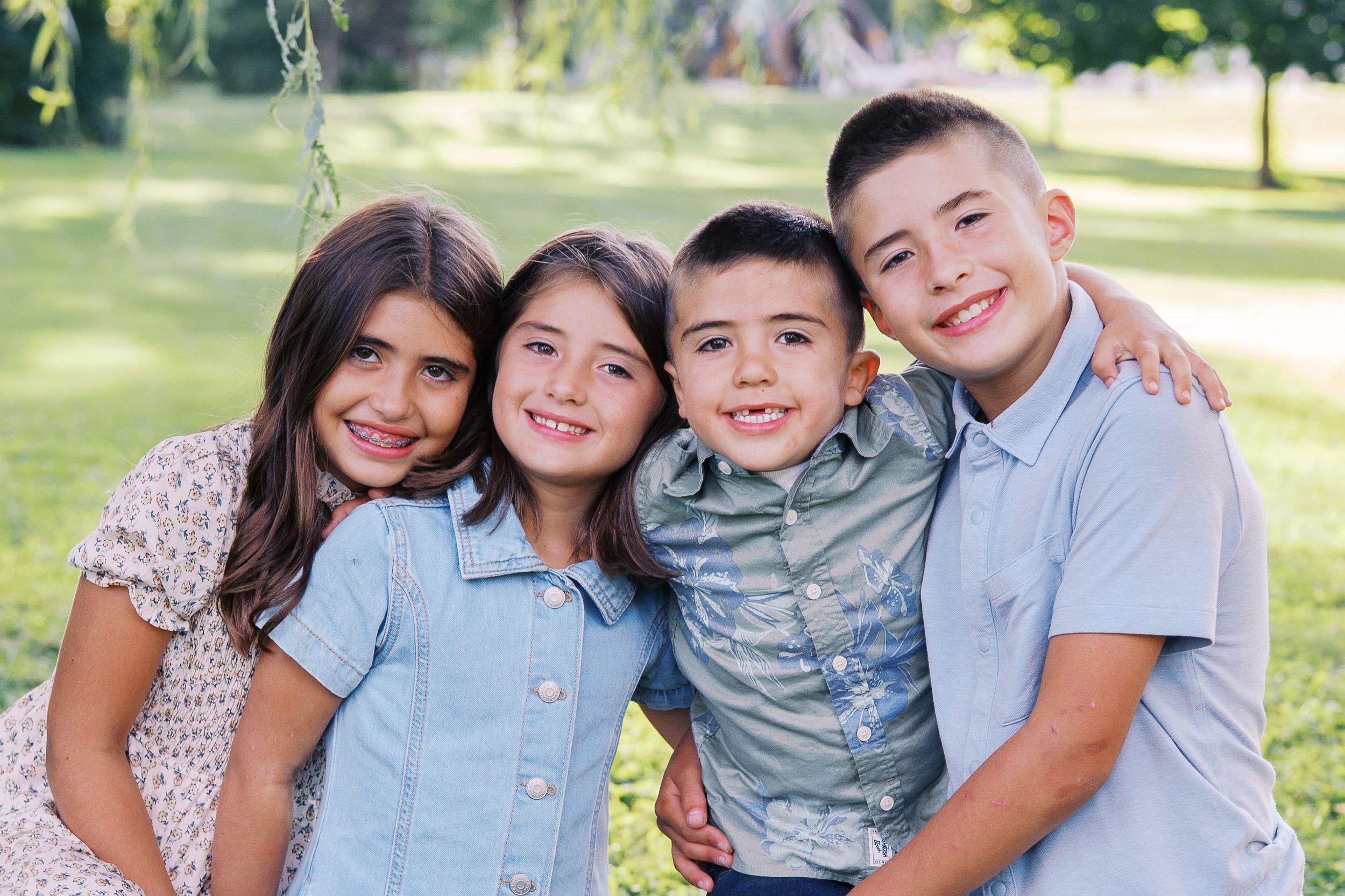 A happy family group poses together outdoors on a sunny day in a grassy park setting.